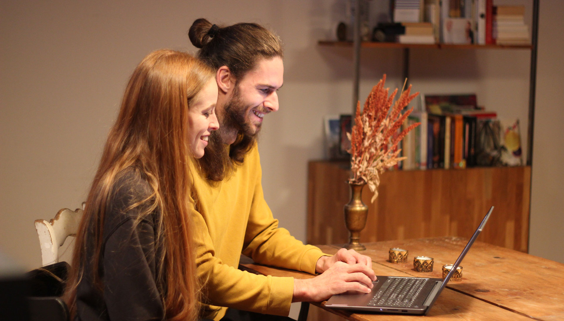 Helen und Dominik sitzen schräg vor einem Laptop und lachen in die Webcam.